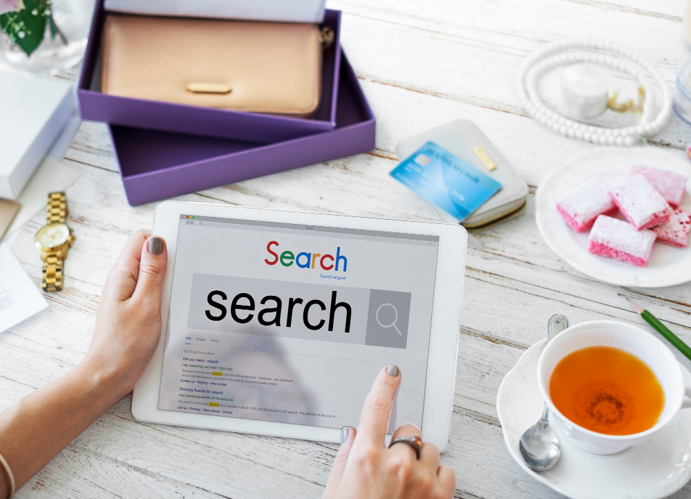 A person using a tablet to search for an SEO keyword, surrounded by a cup of tea, pink sweets, a wallet, and a purple gift box on a white wooden table.