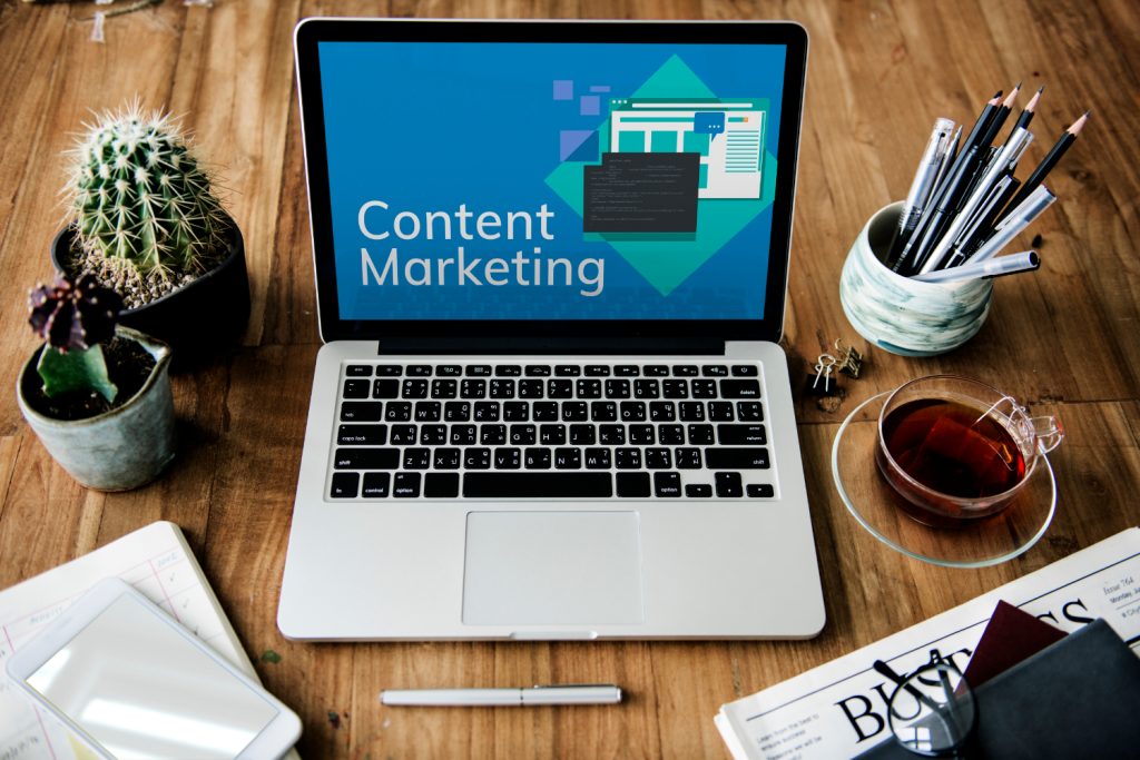 Image of a laptop displaying the text 'Content Marketing' on its screen, surrounded by office essentials like a cactus, pens, a cup of tea, and business documents, symbolizing a workspace dedicated to strategic content marketing efforts.