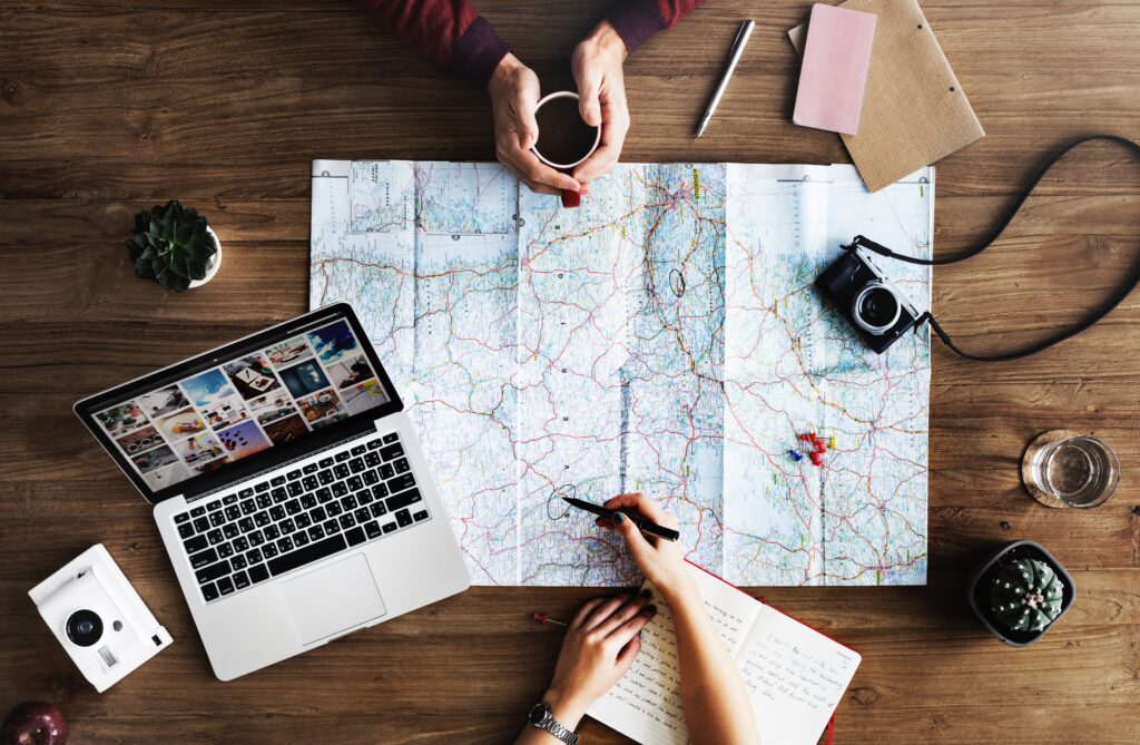 Overhead view of a workspace featuring a map, laptop, camera, and notebook. Two people are engaged in planning, with one writing on the map and the other holding a cup. The laptop screen displays digital media, symbolising creative planning, strategy, and the role of Digital Marketing in travel or project planning.