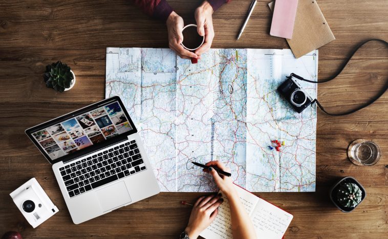 Overhead view of a workspace featuring a map, laptop, camera, and notebook. Two people are engaged in planning, with one writing on the map and the other holding a cup. The laptop screen displays digital media, symbolising creative planning, strategy, and the role of Digital Marketing in travel or project planning.