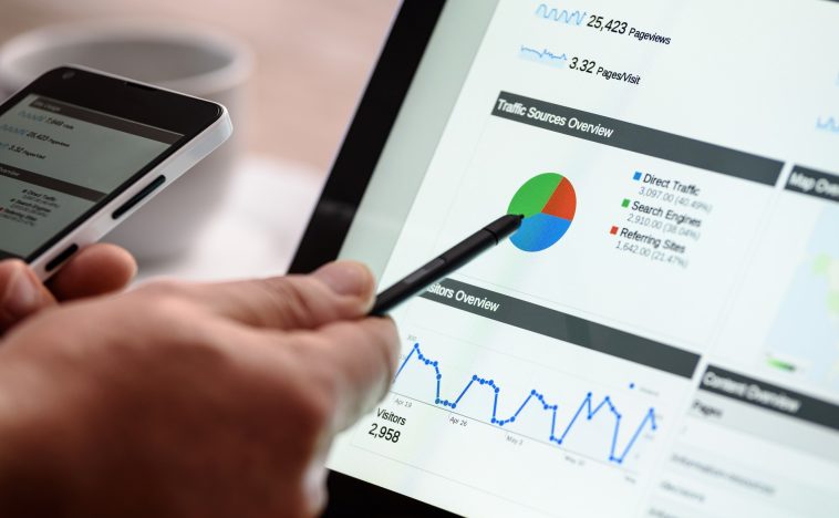 A close-up view of a person holding a smartphone in one hand and using a pen to point at a color-coded pie chart on a laptop screen, highlighting analytics data for effective SEO and Content Marketing.