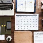 A neatly organized workspace featuring a laptop, calendar, planner, clock, notebook, coffee cups, and a newspaper. The layout emphasizes planning and scheduling, reflecting the importance of strategy and organization in Content Marketing for business growth.