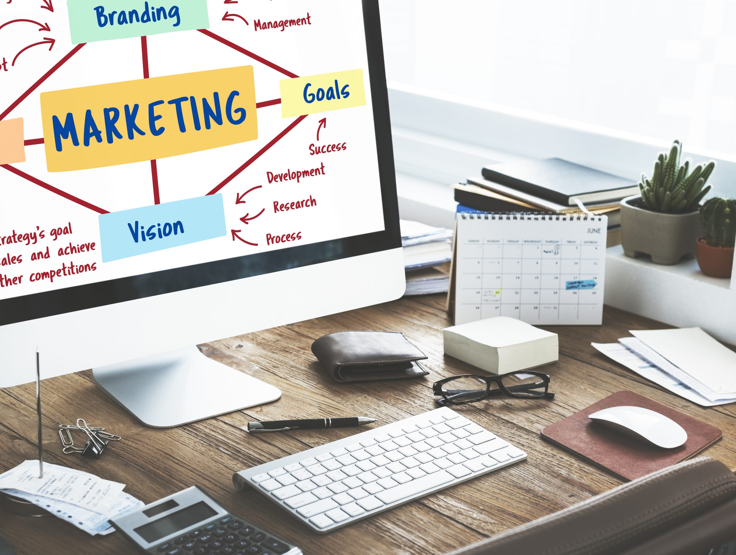 Desktop computer screen displaying a digital marketing strategy diagram with components like branding, goals, vision, and strategy, placed on a wooden office desk with a calendar, glasses, and stationery.