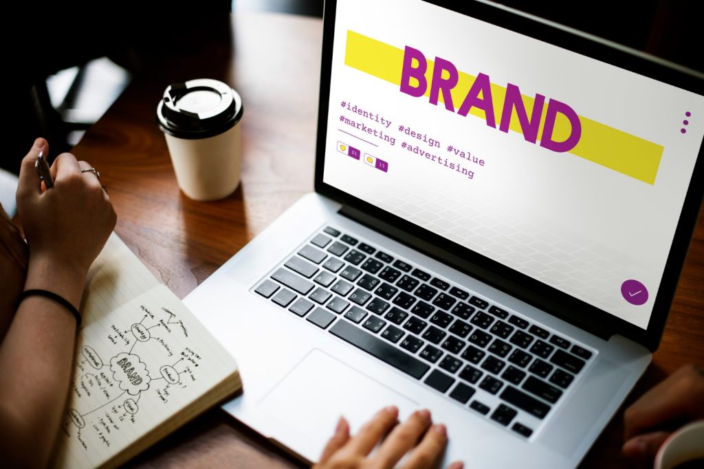 A laptop screen displaying the word 'BRAND' with related hashtags like #identity, #design, #value, and #marketing, symbolizing the concept of brand equity. A person is seen taking notes on branding strategy beside the laptop.