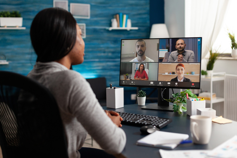 A woman attending a webinar on her desktop computer, with a video conferencing screen displaying four participants in a professional setting.