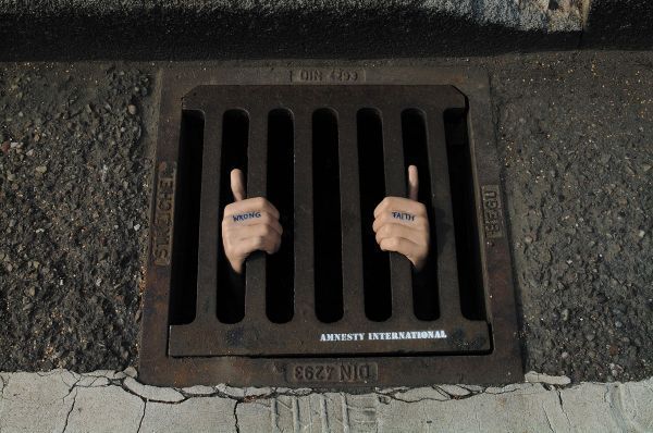 Two hands are visible through a street grate, with the words "WRONG" and "FAITH" tattooed on the knuckles, creating the illusion of a person trapped beneath. This powerful guerrilla marketing ad is by Amnesty International, symbolizing imprisonment for one's beliefs.