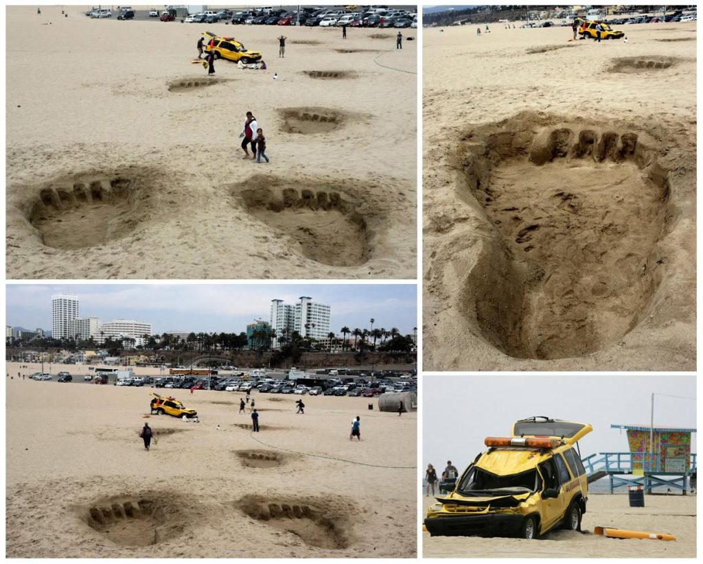 Large footprints are carved into the sand on a beach, leading to a wrecked yellow vehicle. This creative setup gives the impression that a giant creature, like King Kong, walked through the sand and crushed the vehicle as part of a guerrilla marketing campaign.