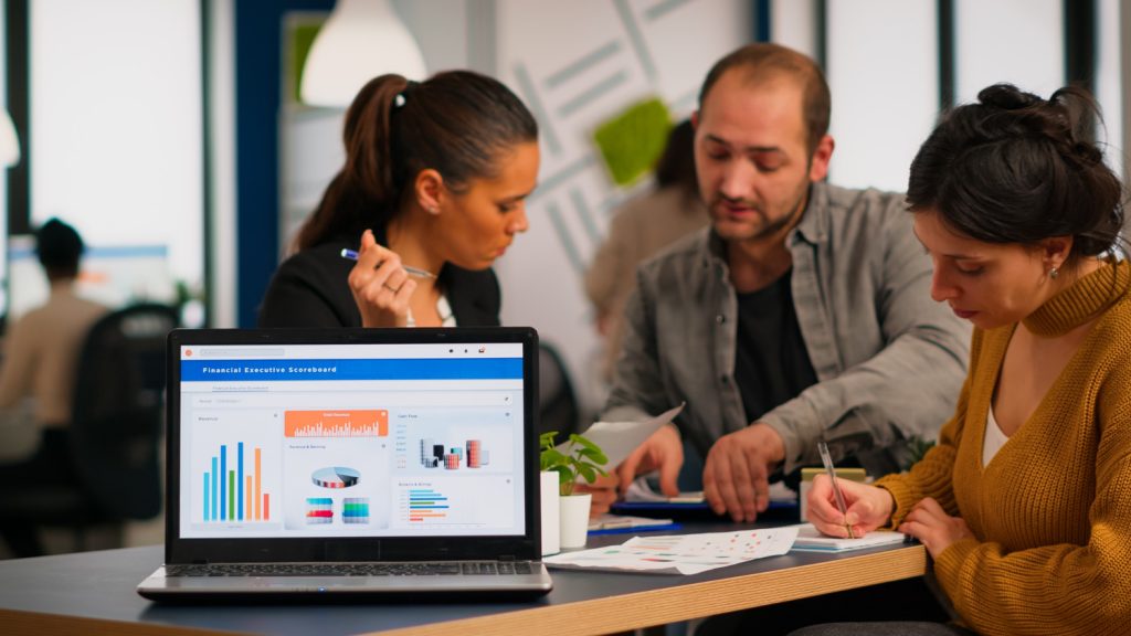 Three professionals collaborating on a B2B marketing strategy, with a laptop screen displaying financial executive scoreboards, charts, and graphs in the foreground.