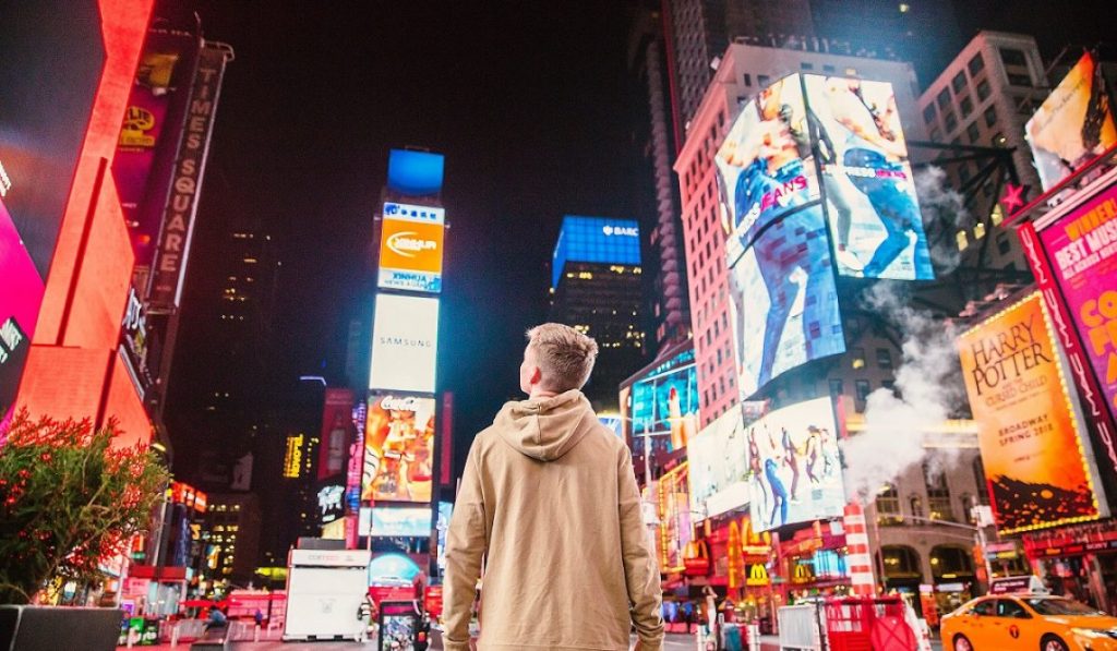 Person standing in a bustling city square surrounded by vibrant billboard advertising, showcasing the impact and reach of outdoor marketing campaigns.