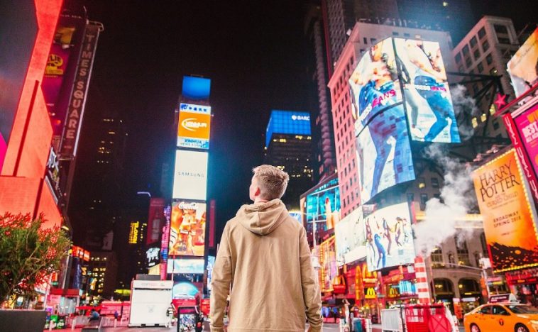 Person standing in a bustling city square surrounded by vibrant billboard advertising, showcasing the impact and reach of outdoor marketing campaigns.