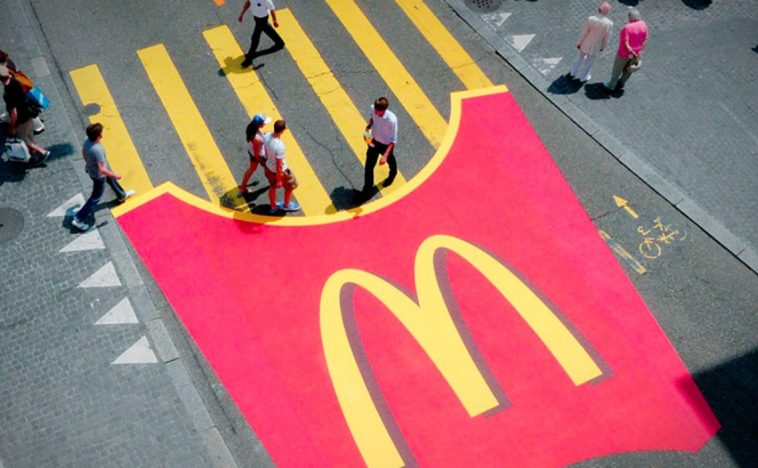 A Guerrilla Marketing advertisement by McDonald's featuring a large red French fries container painted on the pavement, with yellow pedestrian lines representing fries. Pedestrians are seen walking across the lines.